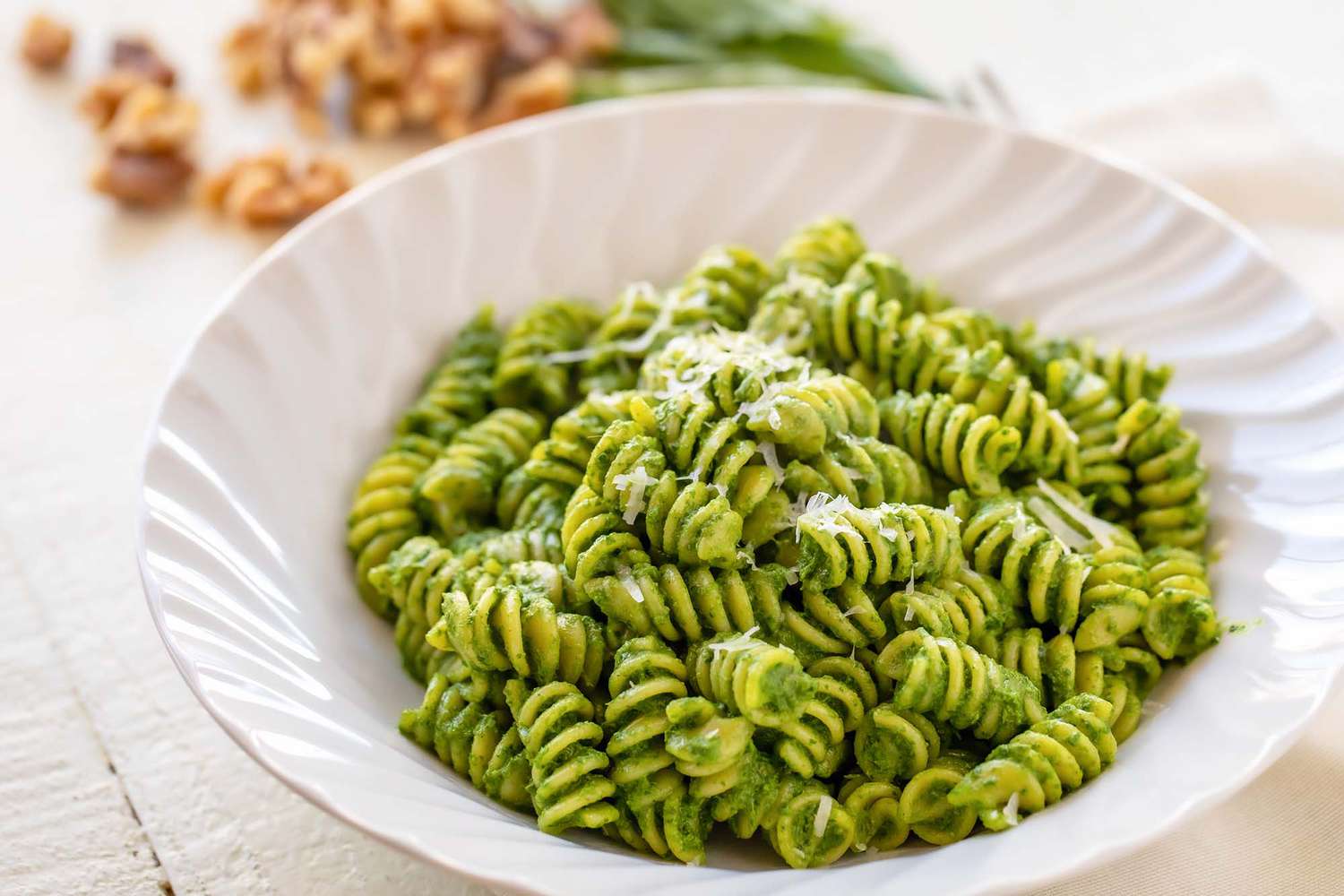 Horizontal view of a white bowl with rotini and kale pesto inside. Walnuts and basil are in the background.