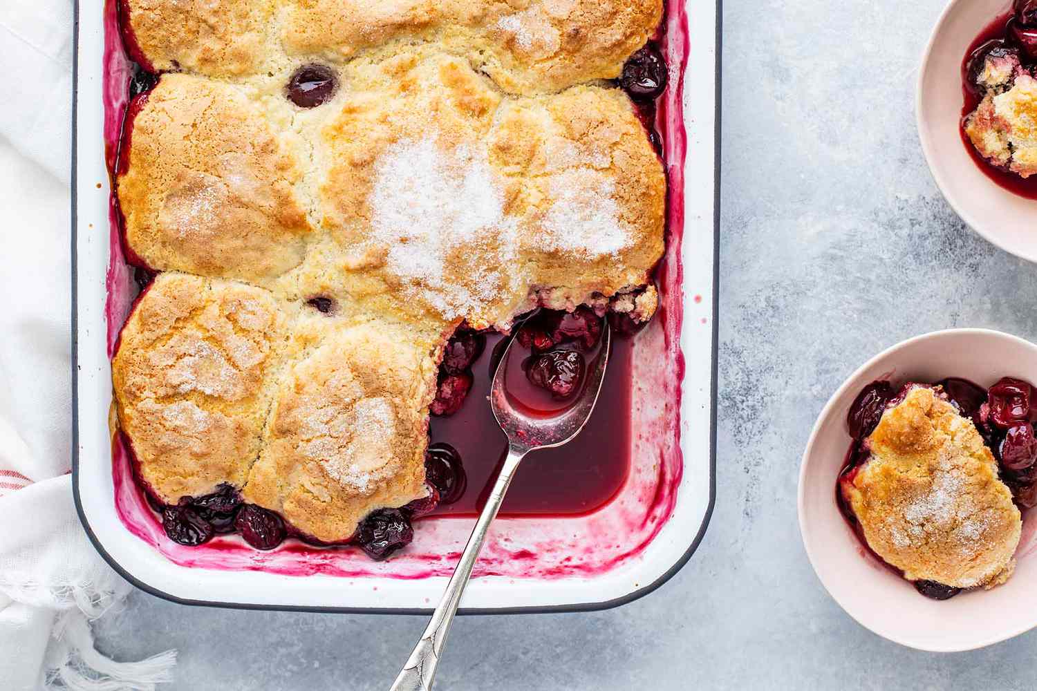 Cherry cobbler in a white dish with a spoon and two servings in bowls on the side