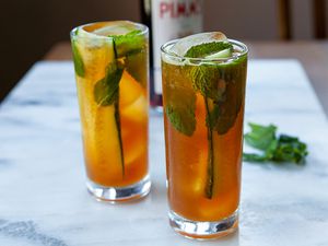 Two tall glasses of Pimms cup cocktail with slices of cucumber, mint leaves and ice. Mint leaves and Pimms no 1 are behind the glasses.