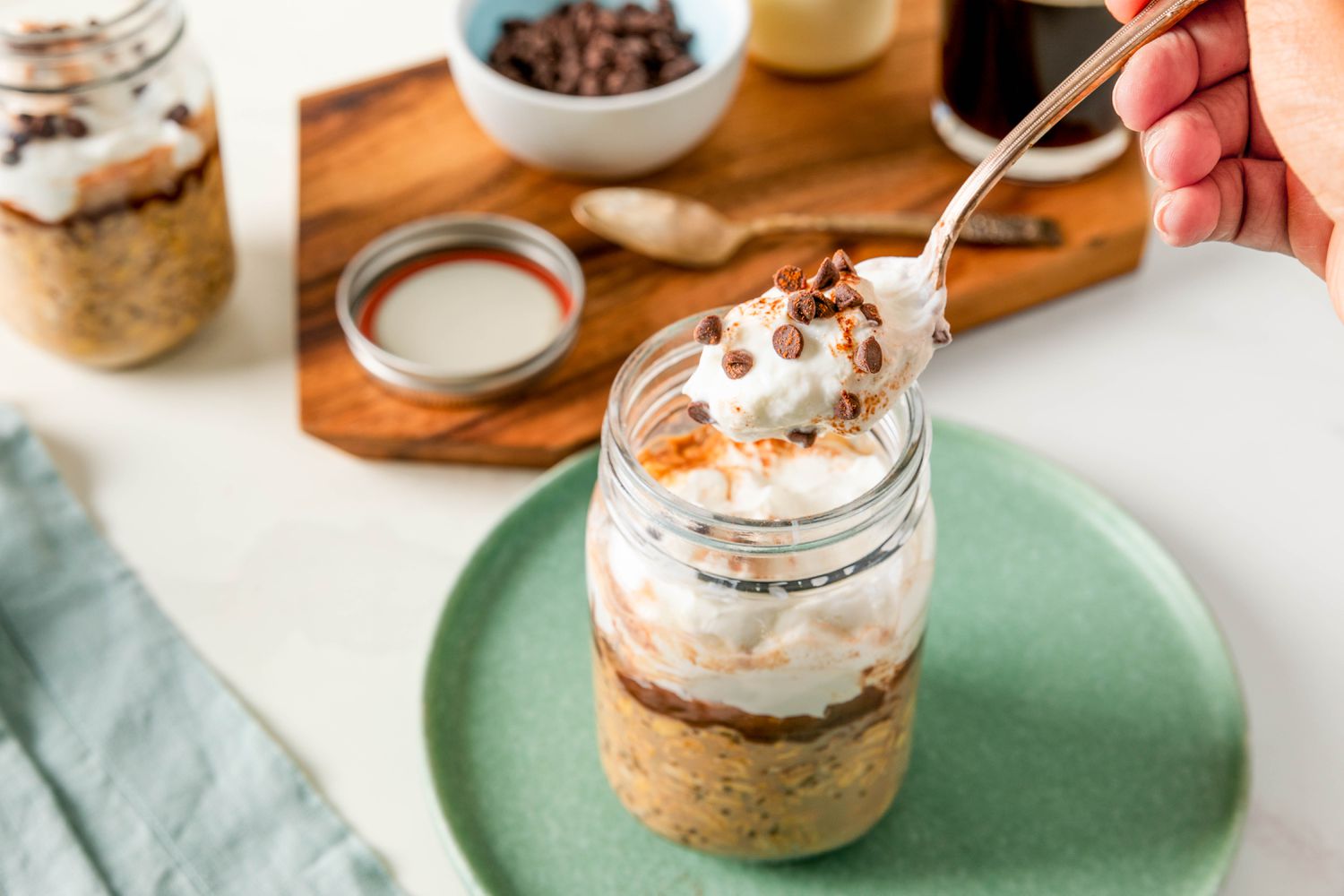 Spoonful of tiramisu overnight oats lifted from a jar on a small plate, and in the surroundings, another jar of oats and a robin egg colored kitchen towel on the counter and on a wooden board, the jar lid, a small bowl of mini chocolate chips and a glass of coffee