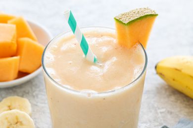 Glassful of Cantaloupe Smoothie With a Slice of Cantaloupe on the Glass Rim, and in the Surroundings, a Bowl of Cantaloupe and Slices of Bananas and a Whole Banana on the Counter