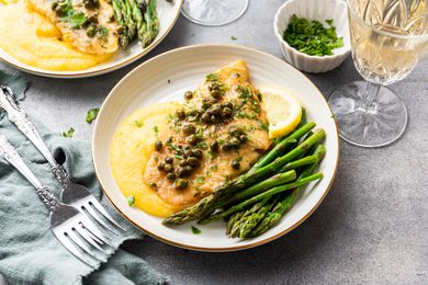 Sole Piccata with Asparagus, Polenta, and Lemon Slice Topped with Capers Surrounded by Another Plate of Sole Piccata, Forks, a Glass, and a Bowl of Herbs 