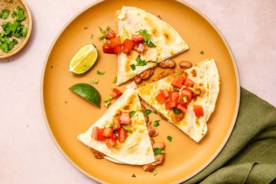 Three slices of a quesadilla on a plate along with lime slices at a table setting with a table napkin and small plate of cilantro 