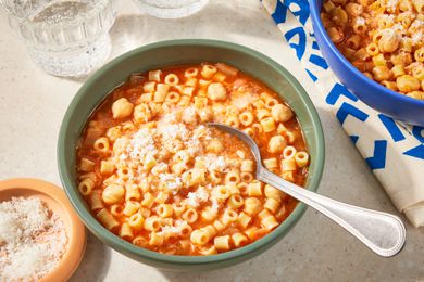 Overhead view of a green bowl of Pasta e Ceci and a spoon topped with parmesan cheese next to a bowl of parmesan, another bowl of pasta and a glass of carbonated beverage
