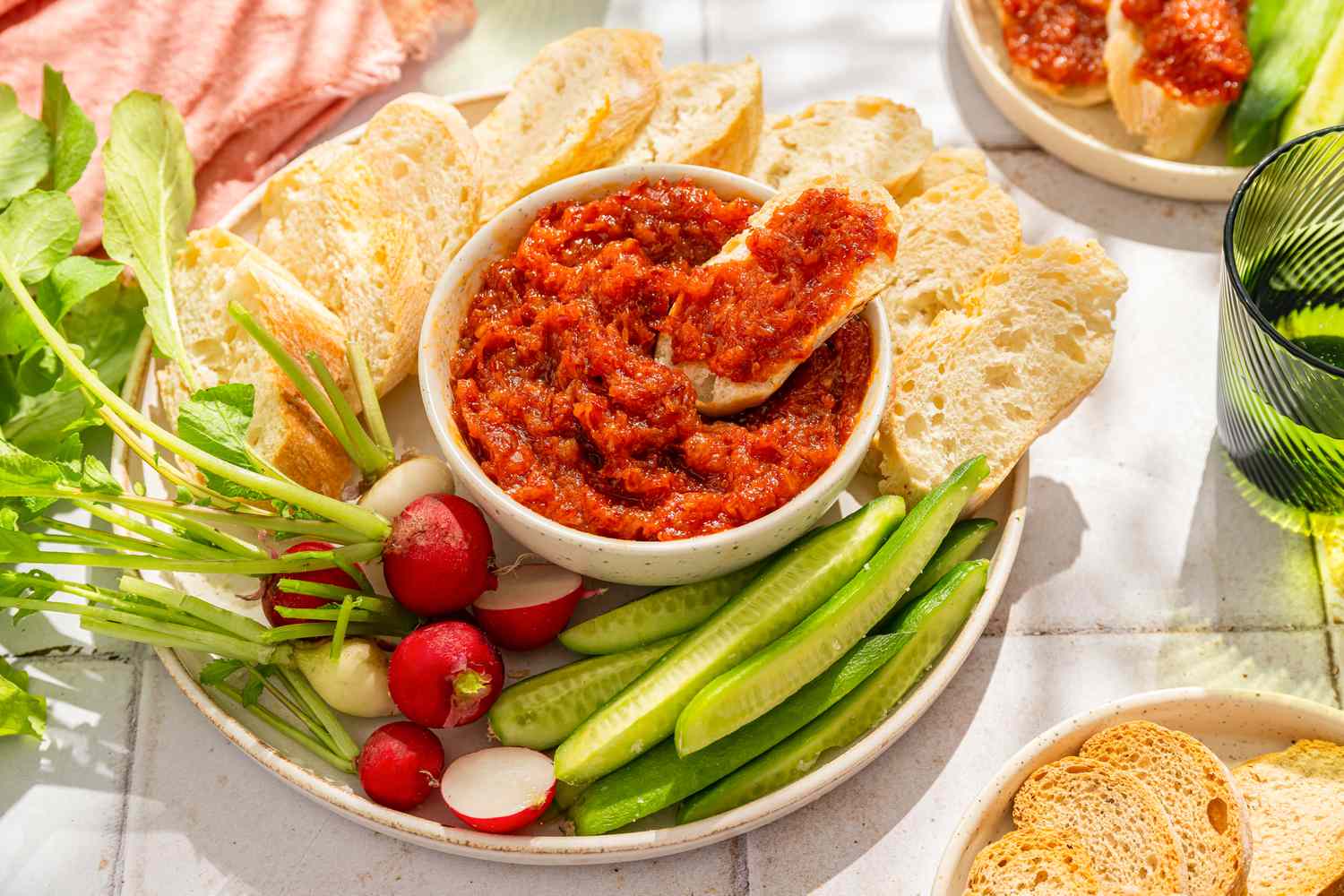 Side view of 2-ingredient honey chorizo spread in a bowl surrounded by radishes, cucumber spears and sliced baguette, with one baguette slice topped with the spread