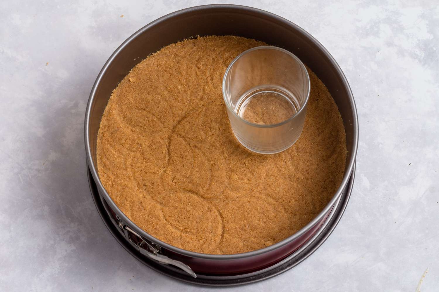 Using a cup to press the graham cracker crust into the springform pan to make an easy no bake cheesecake recipe.