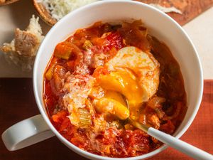 Overhead view of Acquacotta (Tuscan Soup) in a white bowl with a spoon plus bread on a cutting board and Pecorino romano cheese in a bowl to the side
