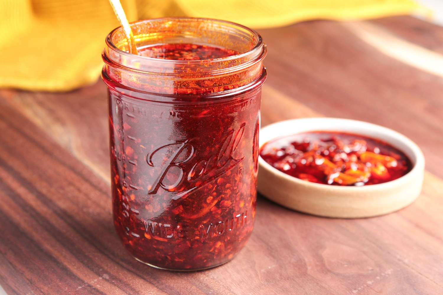 Mason Jar Filled With Chili Crisp, and in the Background, a Saucer With Some Homemade Chili Crisp and a Mustard Kitchen Towel