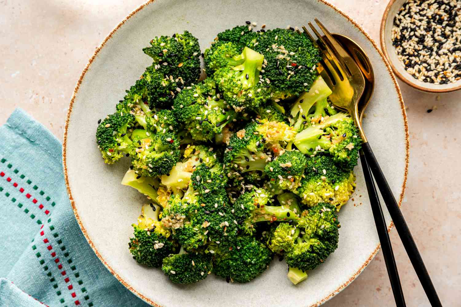 Bowl of Microwave Broccoli with Sesame Dressing with Utensils, Next to a Table Napkin, a Glass of Water, and a Bowl with More Sesame Seeds