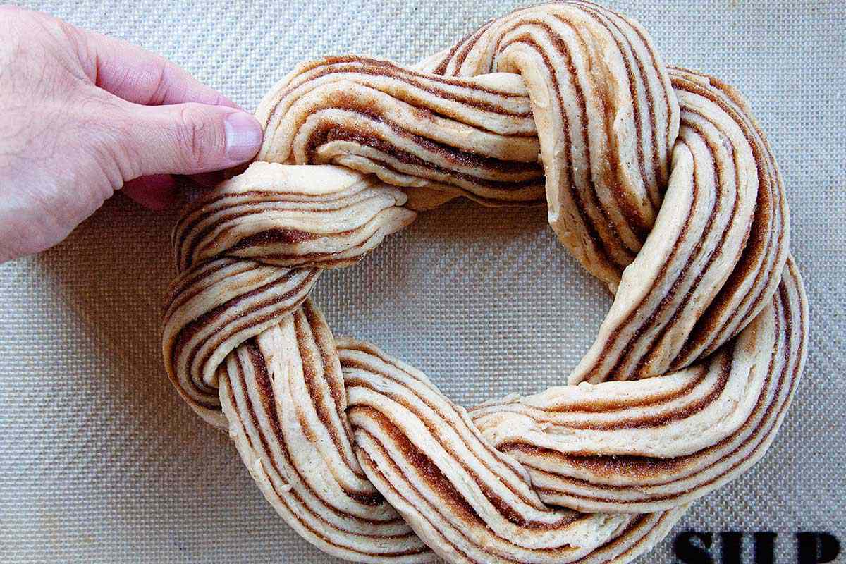 The twisted, circular King Cake dough is on a baking sheet. A hand is tucking the ends under to hide them.