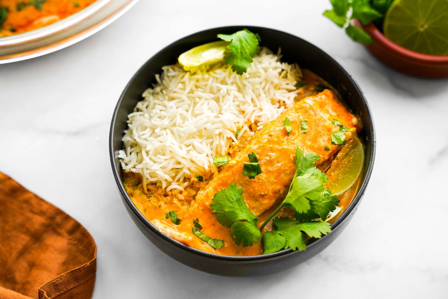 Bowl of Microwave Red Curry Poached Salmon with Rice and Garnished with Cilantro and Lime Wedge, and in the Surroundings, a Kitchen Towel, a Bowl with More Curry, and a Bowl with a Halved Lime and Cilantro