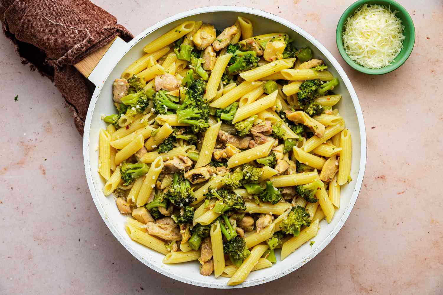 Pan of Chicken and Broccoli Pasta with the Handle Wrapped in a Kitchen Towel and Next to It, a Small Bowl of Shredded Cheese