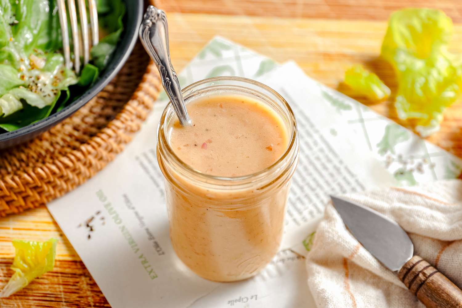A jar of creamy dressing with a spoon placed on a napkin and a wooden surface with salad and a knife nearby