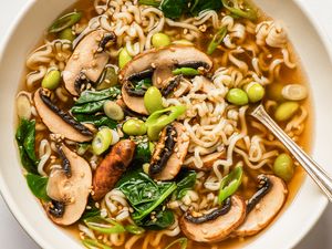 A bowl of noodle soup containing mushrooms spinach edamame green onions and sesame seeds with a spoon on the side