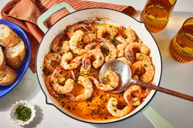 spanish garlic shrimp in a dutch oven at a table setting with forks on a table napkin, glasses of beer, and a serving of spanish garlic shrimp with baguette slices in a bowl