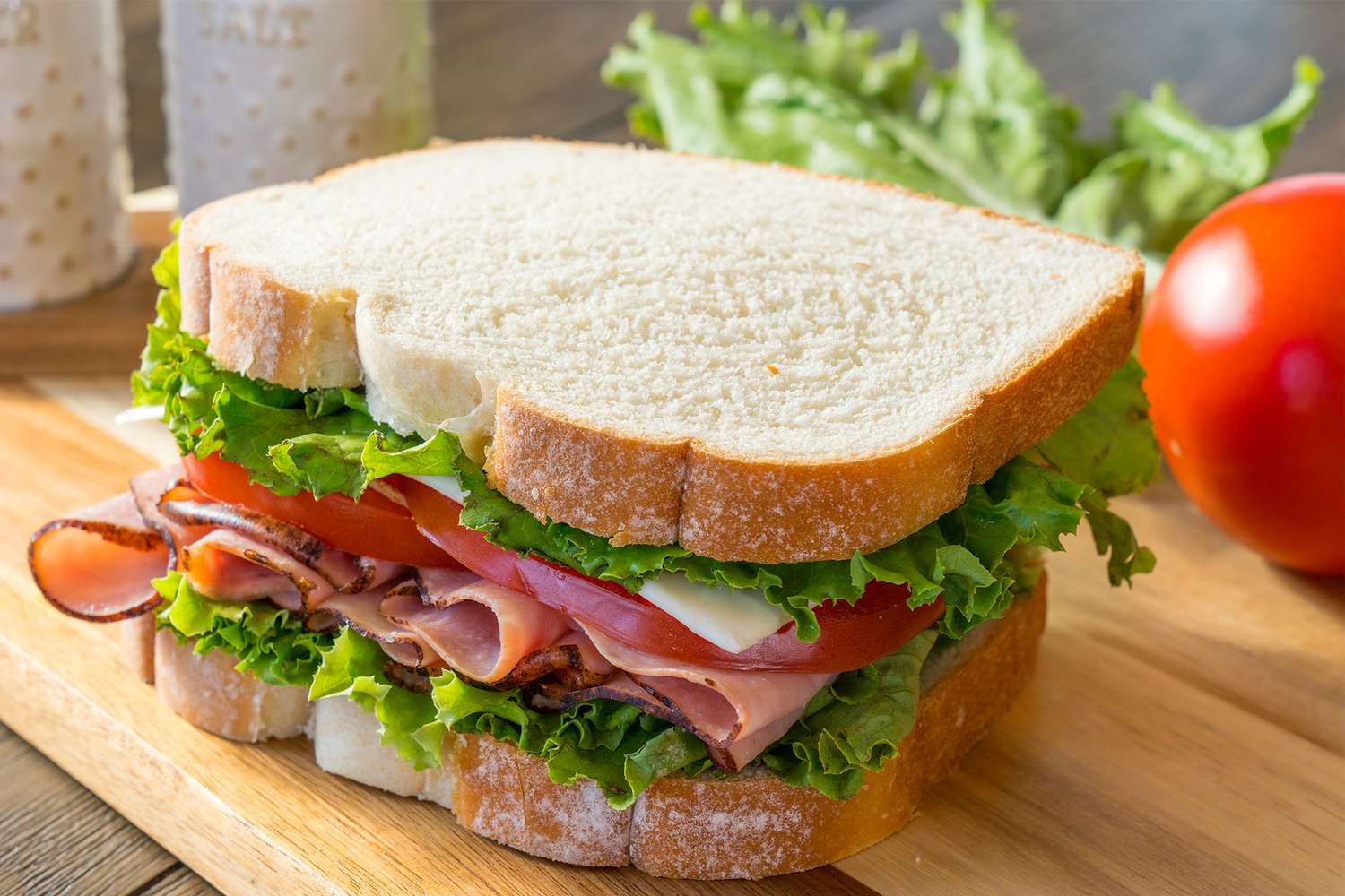 A sandwich containing stacked deli meats lettuce tomato and cheese on sliced bread placed on a wooden surface with a tomato in the background