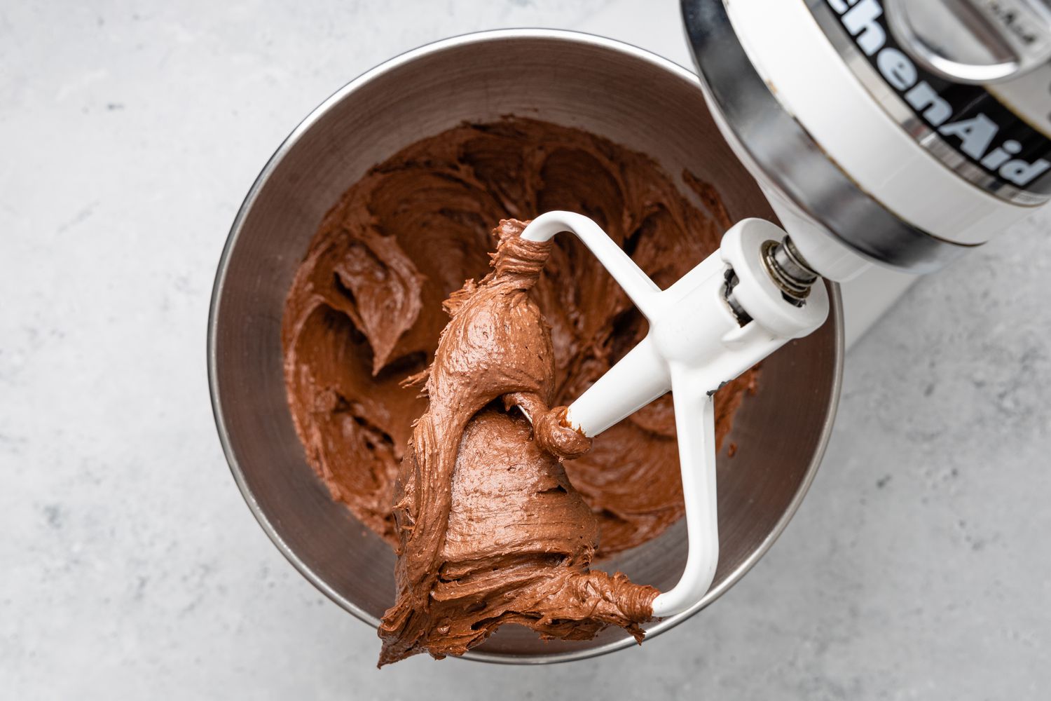 Chocolate Buttercream Frosting on a Mixer Paddle Attachment With the Rest in the Bowl Below It
