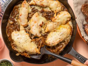 french onion chicken in a skillet at a table setting with glasses, a bowl of bread, and a small bowl of herbs