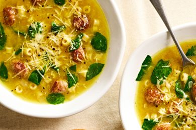 Two bowls of soup with pasta spinach meatballs and grated cheese topped with ground pepper