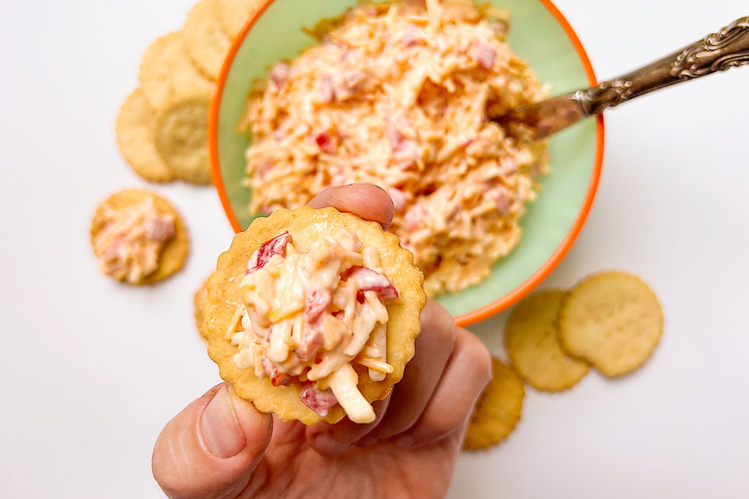 a hand holding up a ritz cracker with pimento cheese and in the background, a bowl of pimento cheese and more crackers on the counter 