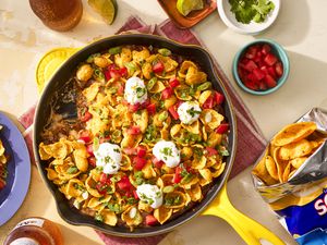 Frito pie dip in a cast iron skillet at a table setting with a bag of fritos lays, a single serving of dip on a plate, a small bowl of diced tomatoes, and a beer