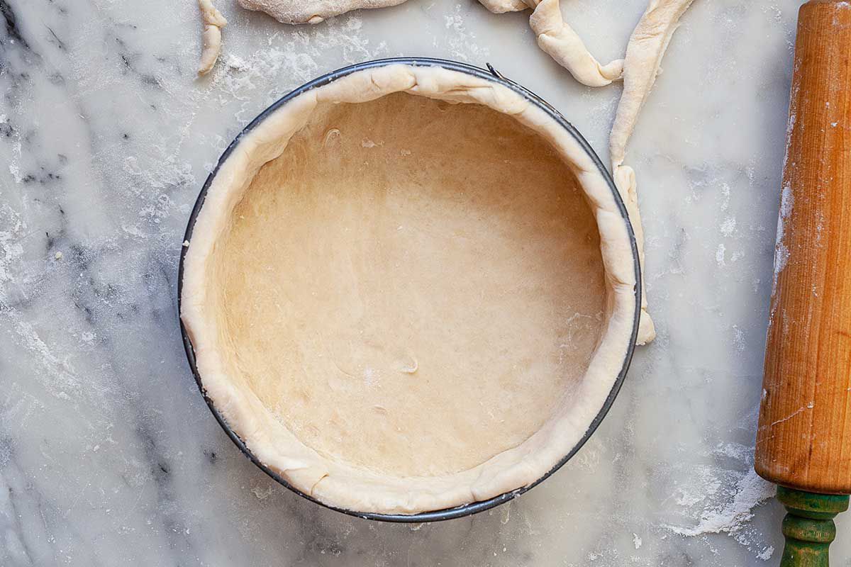 A springform pan has an unbaked quiche crust inside. Scraps of dough and a wooden cutting board are to the right of the pan.