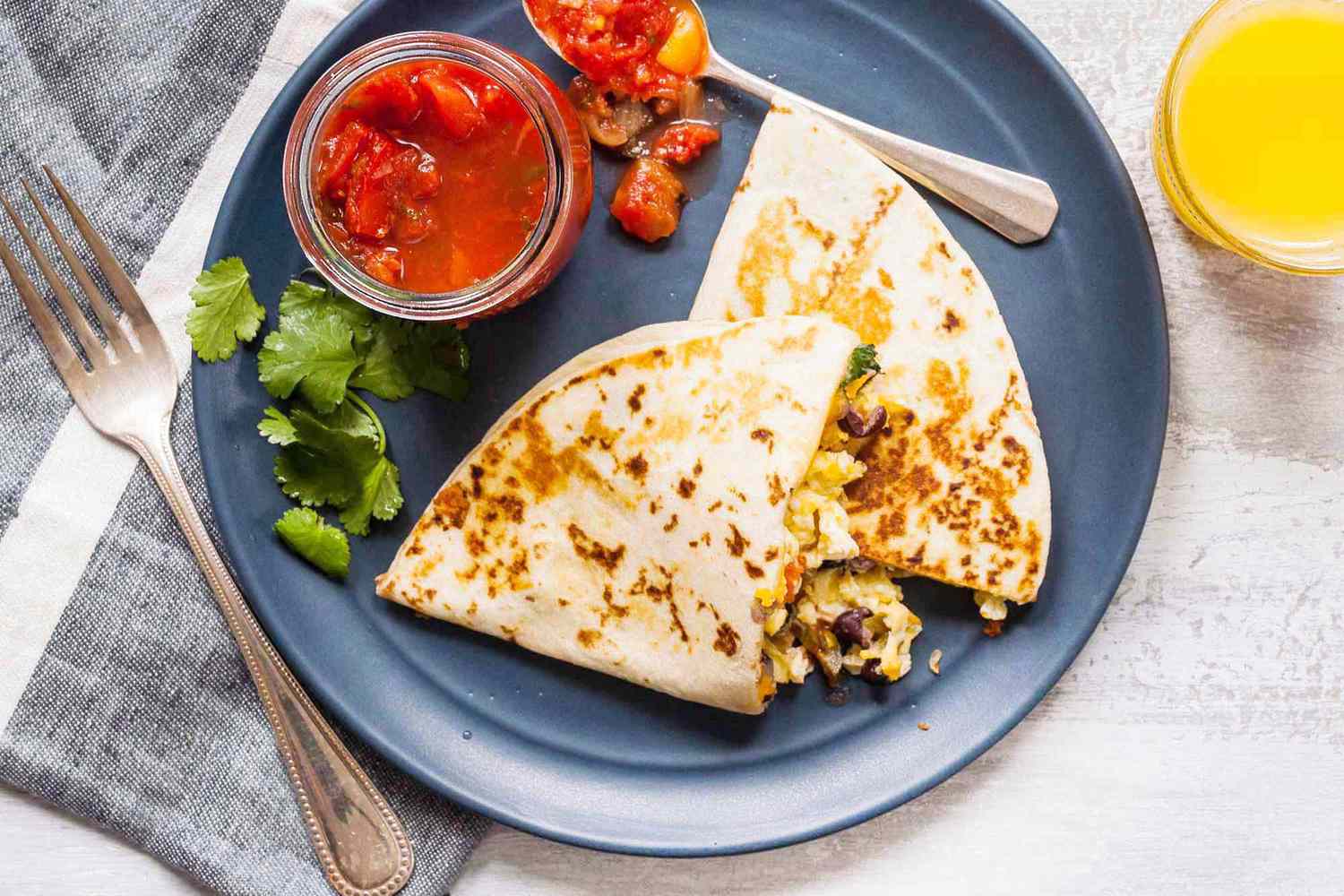 A blue plate with two freezer-friendly breakfast quesadilla triangles, cilantro, salsa and a spoon with salsa on the plate. A glass of orange juice is to the right of the plate. A fork and grey linen are to the left of the plate.