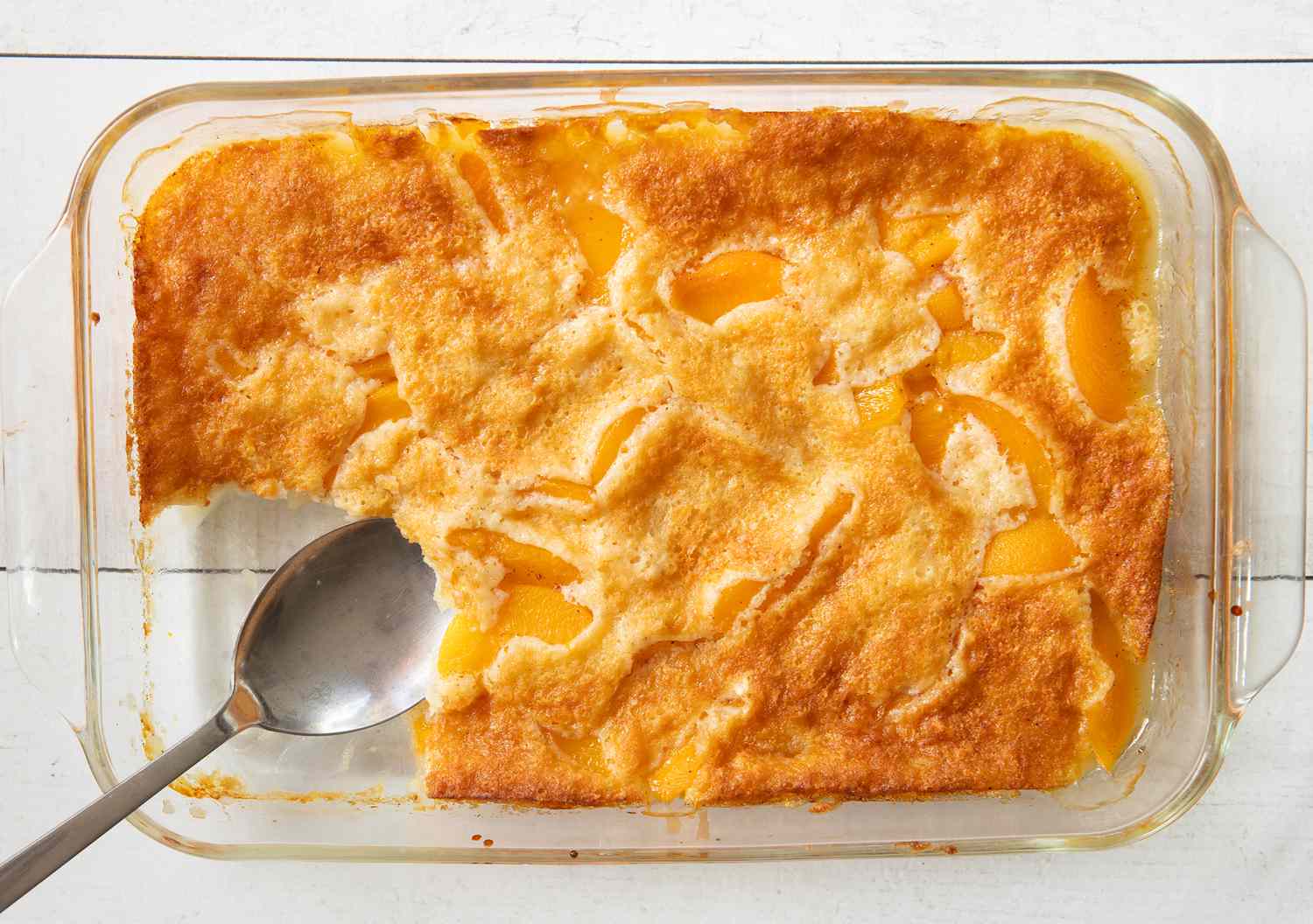 Baking dish containing peach cobbler with a serving spoon and a portion removed