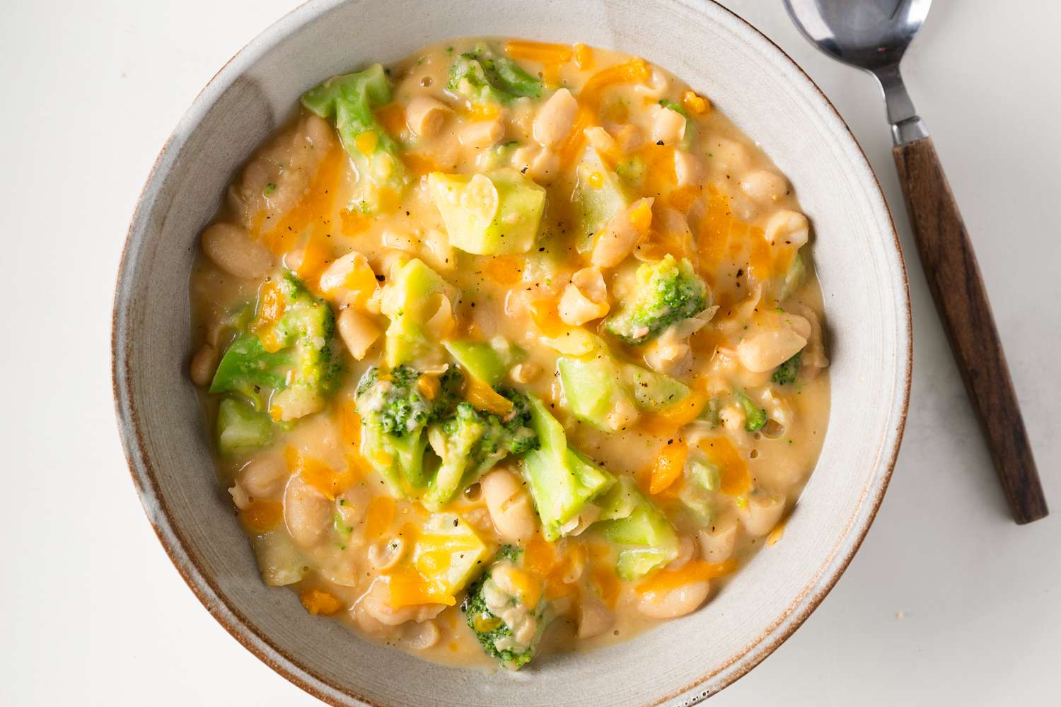 Overhead view of a stoneware bowl of Broccoli Cheddar Creamy Beans next to a spoon and all on a white background