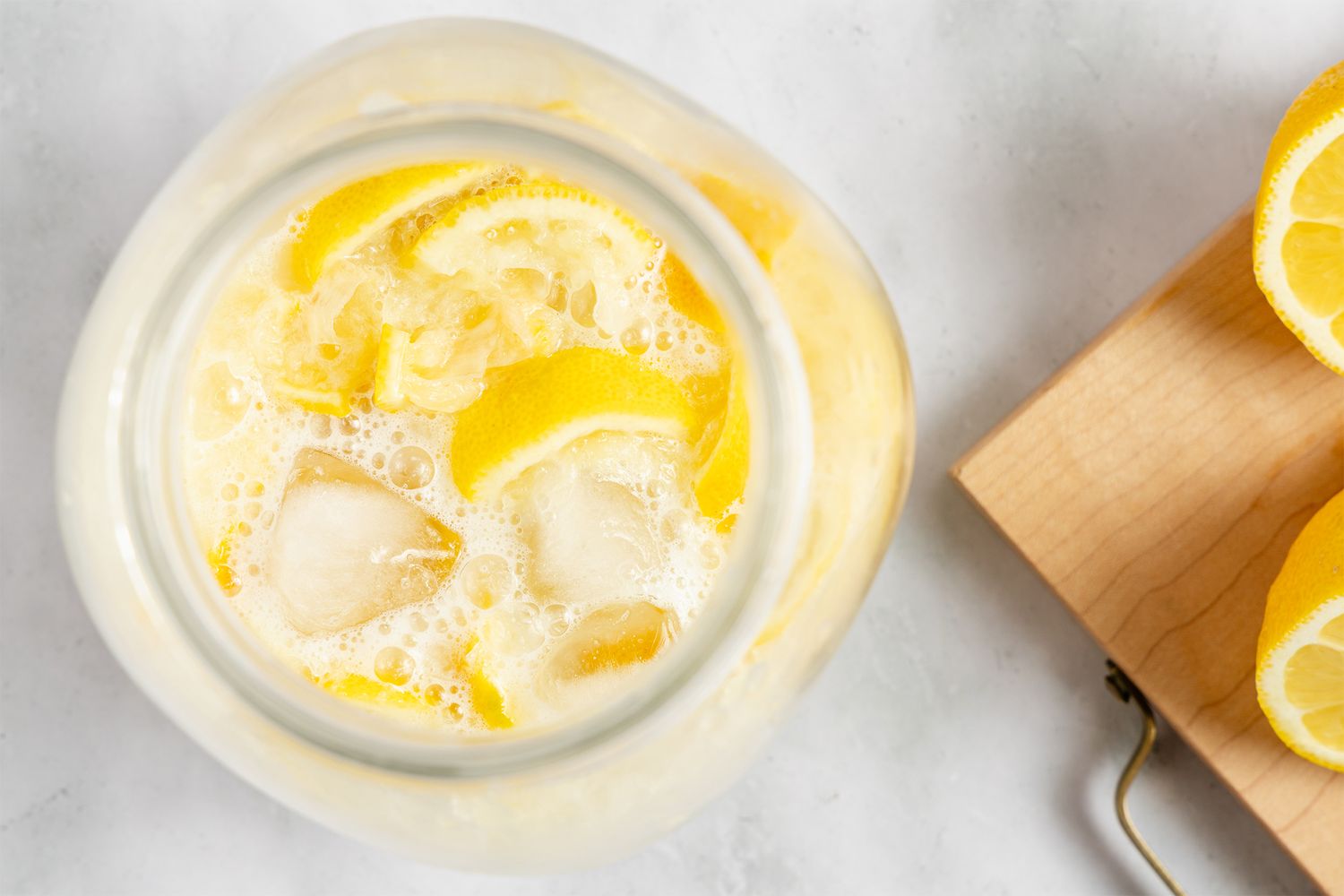 A jar of creamy lemonade with slices of lemon and ice on a table next to lemon halves on a cutting board