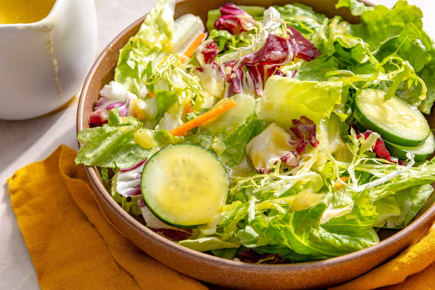 Angled view of a brown bowl of lettuce and cucumbers topped with Miso-Maple Dressing all on a tan countertop