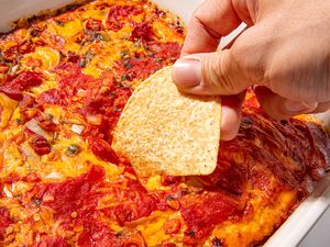 Hand dipping a tortilla chip into a baked tomato and cheese dip