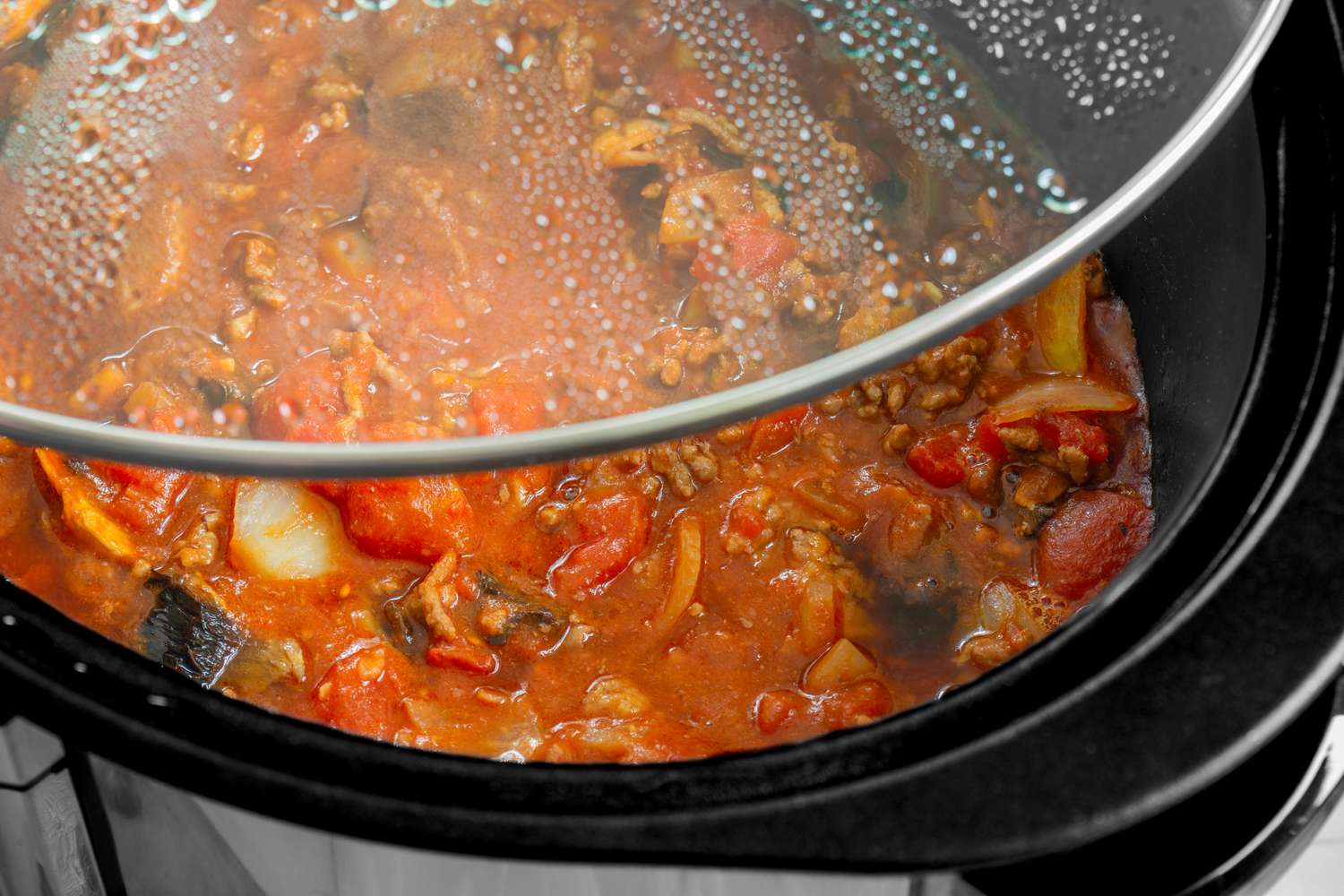A slow cooker with a tomatobased stew visible through a glass lid containing meat onions and other vegetables