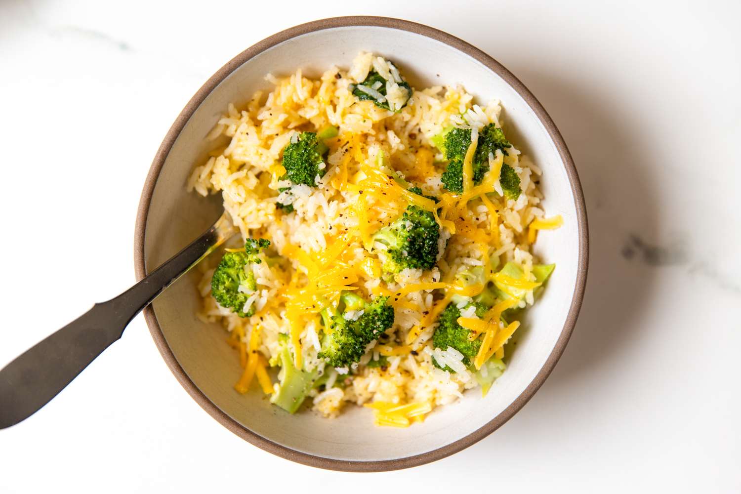 A bowl of rice mixed with broccoli and topped with melted cheese a spoon on the side