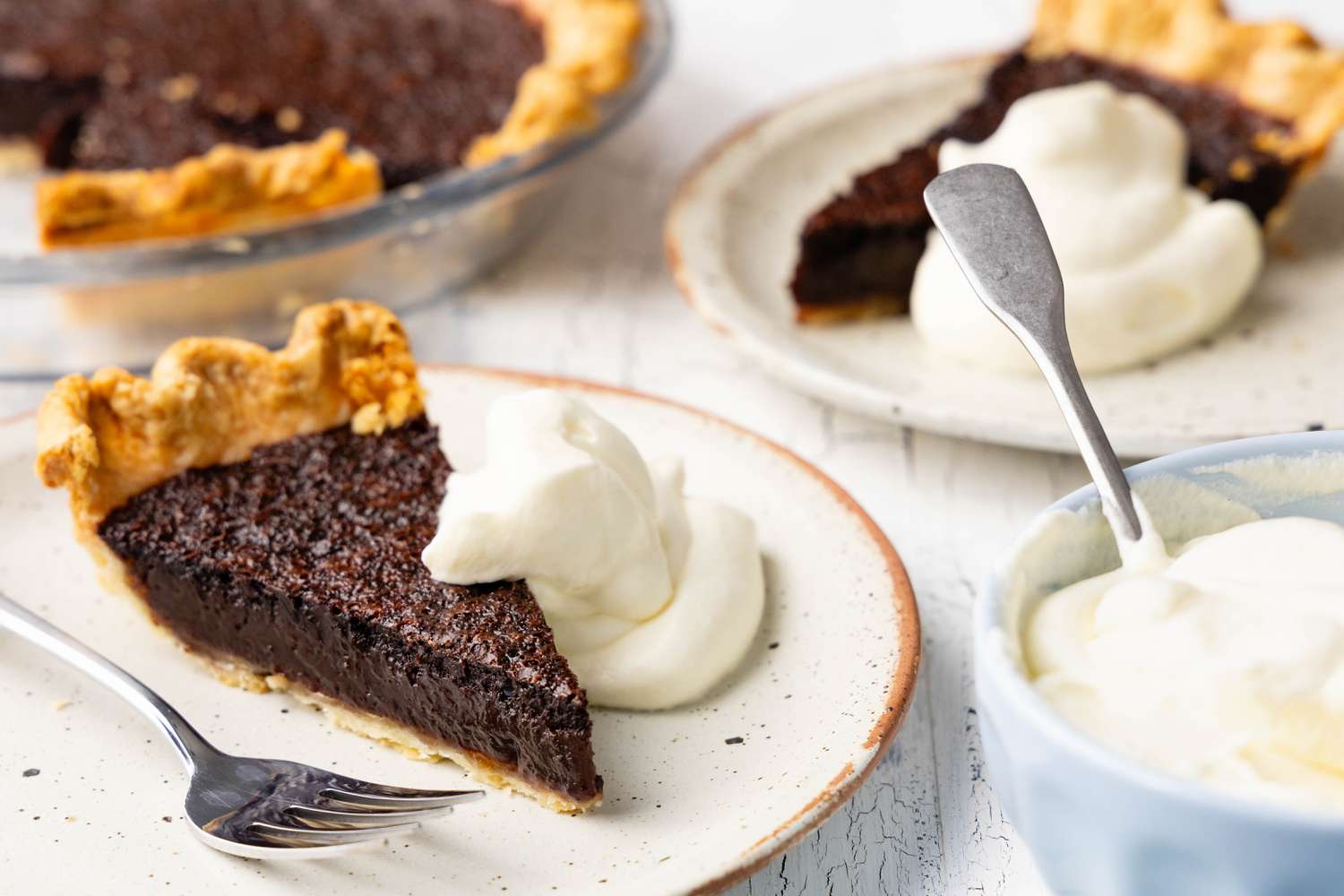 Slice of Chocolate Chess Pie Served With a Dollop of Cream on the Side (on a Plate), and in the Background, More Plates With Slices of Pie and Whipped Cream, a Bowl With More Whipped Cream, and a Pie Dish With More Chocolate Chess Pie 