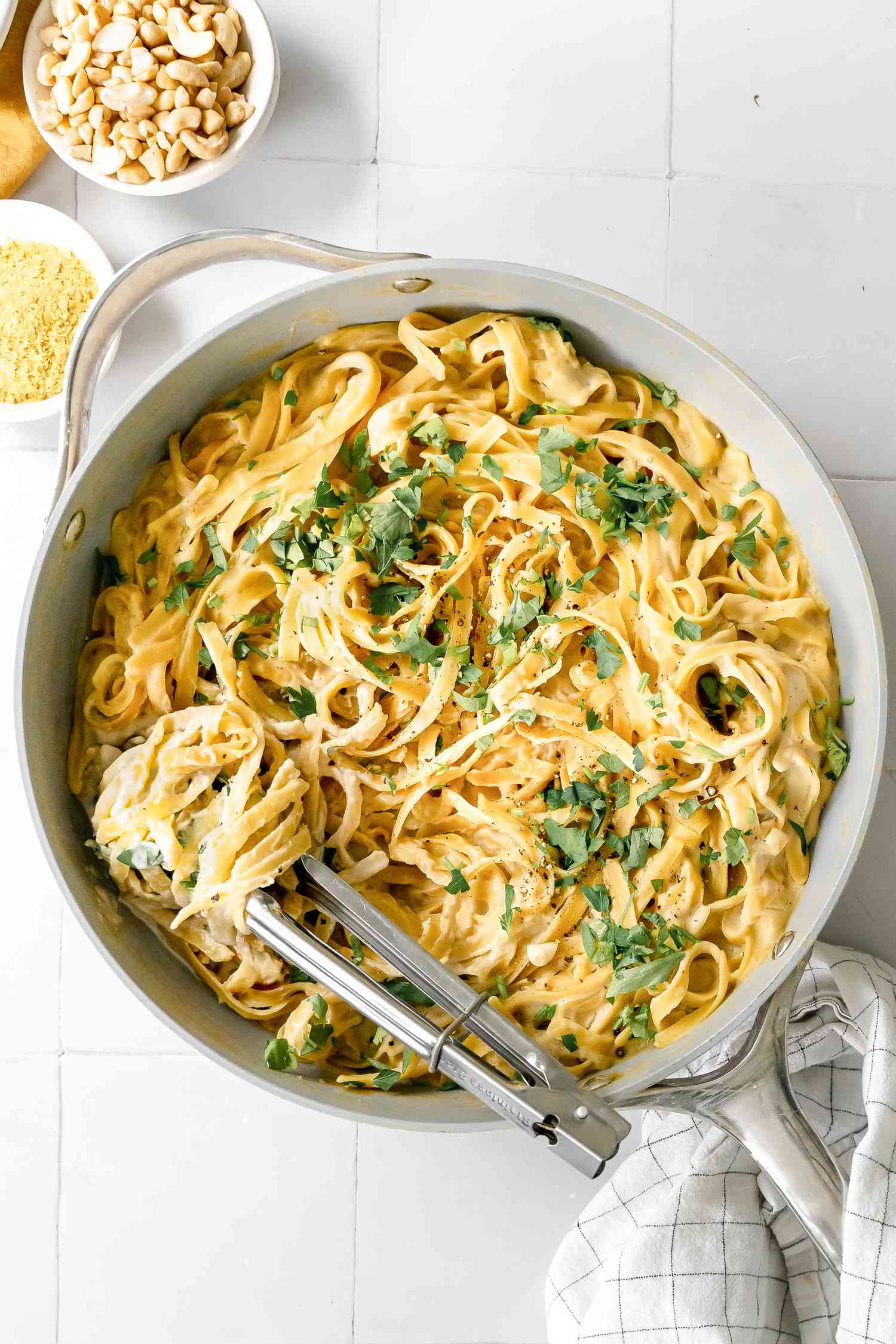 Pan of Super Creamy Vegan Pasta Topped with Parsley and Some Wrapped Around a Pair of Tongs. Next to the Pan, Two Bowls of Ingredients (One: Cashews and Two: Nutritional Yeast)