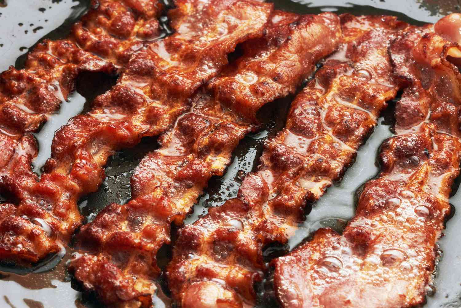 Deeply browned and crisp-looking slices of bacon cooking in a cast iron skillet