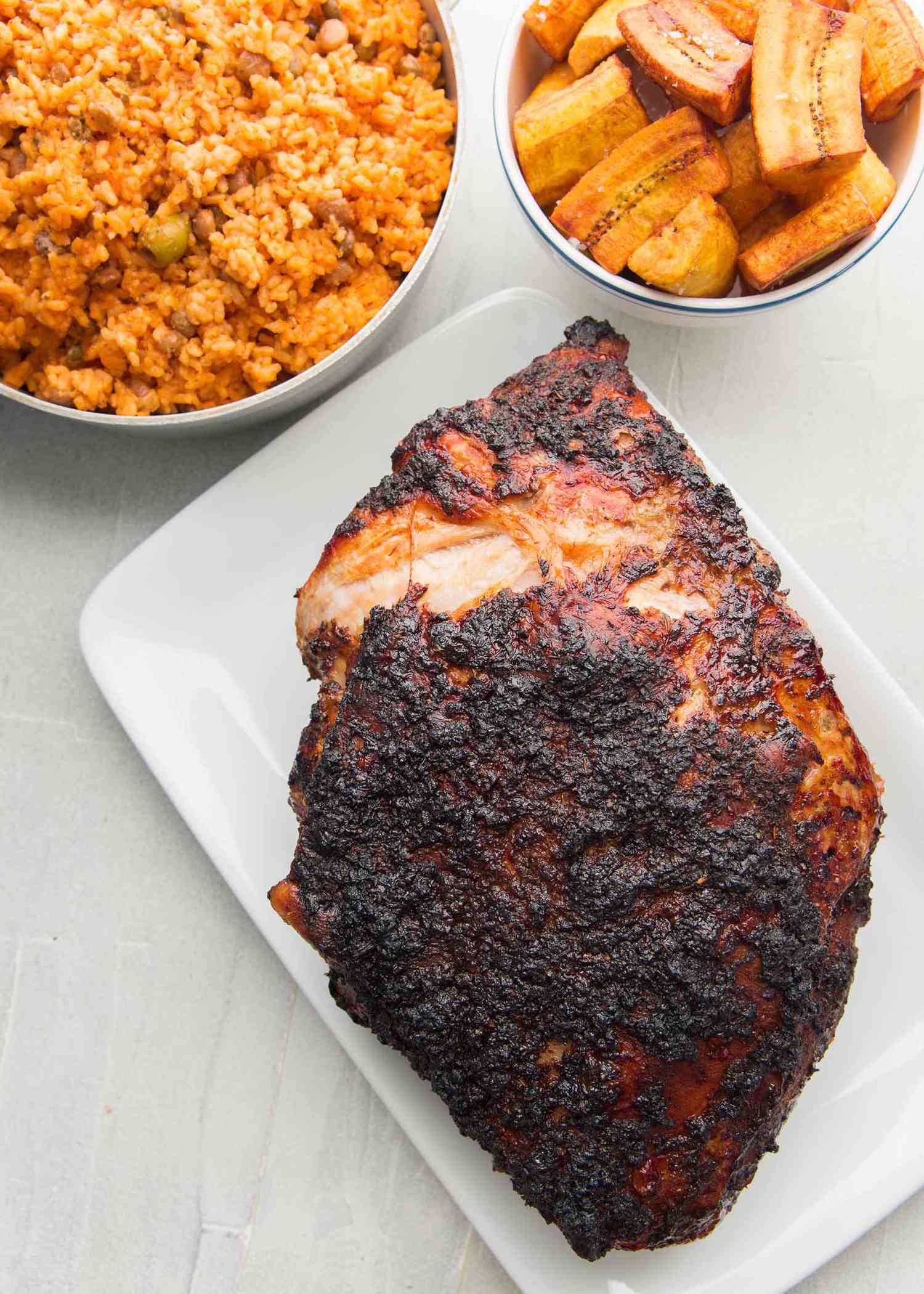 Top view of mojo marinated pork roast with a dark crust sits on a white platter. A bowl of fried plantains and a bowl of red rice are above the pork.