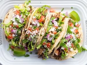 Four tacos filled with shredded pork, lettuce, diced tomatoes, crumbled cheese, and onions on a plate