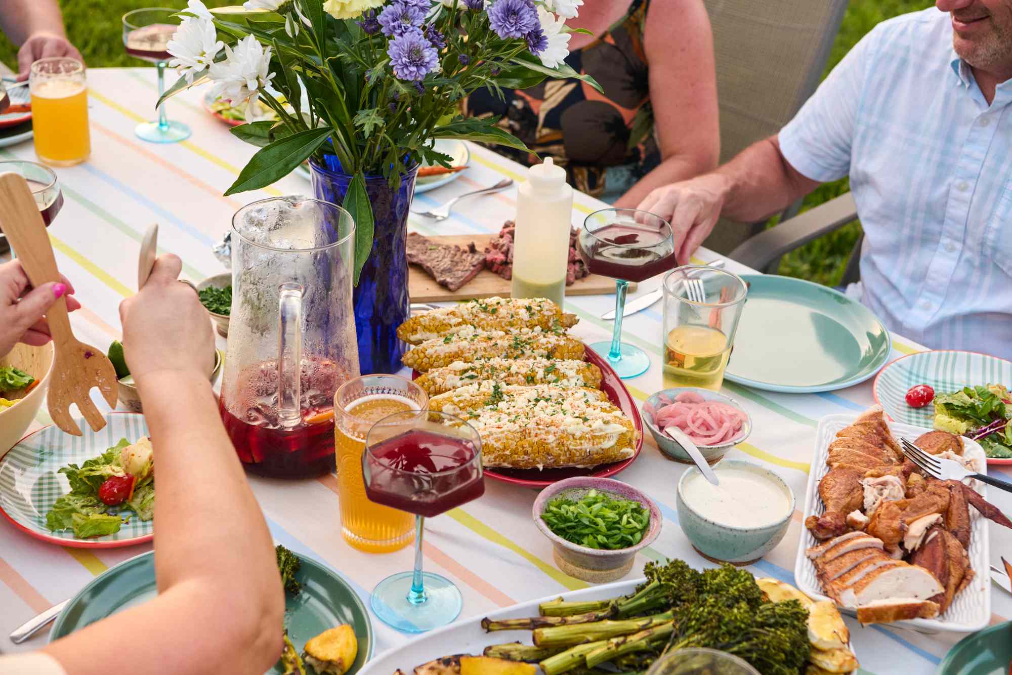 Outdoor table set with various dishes, drinks, and surrounded by people enjoying a meal