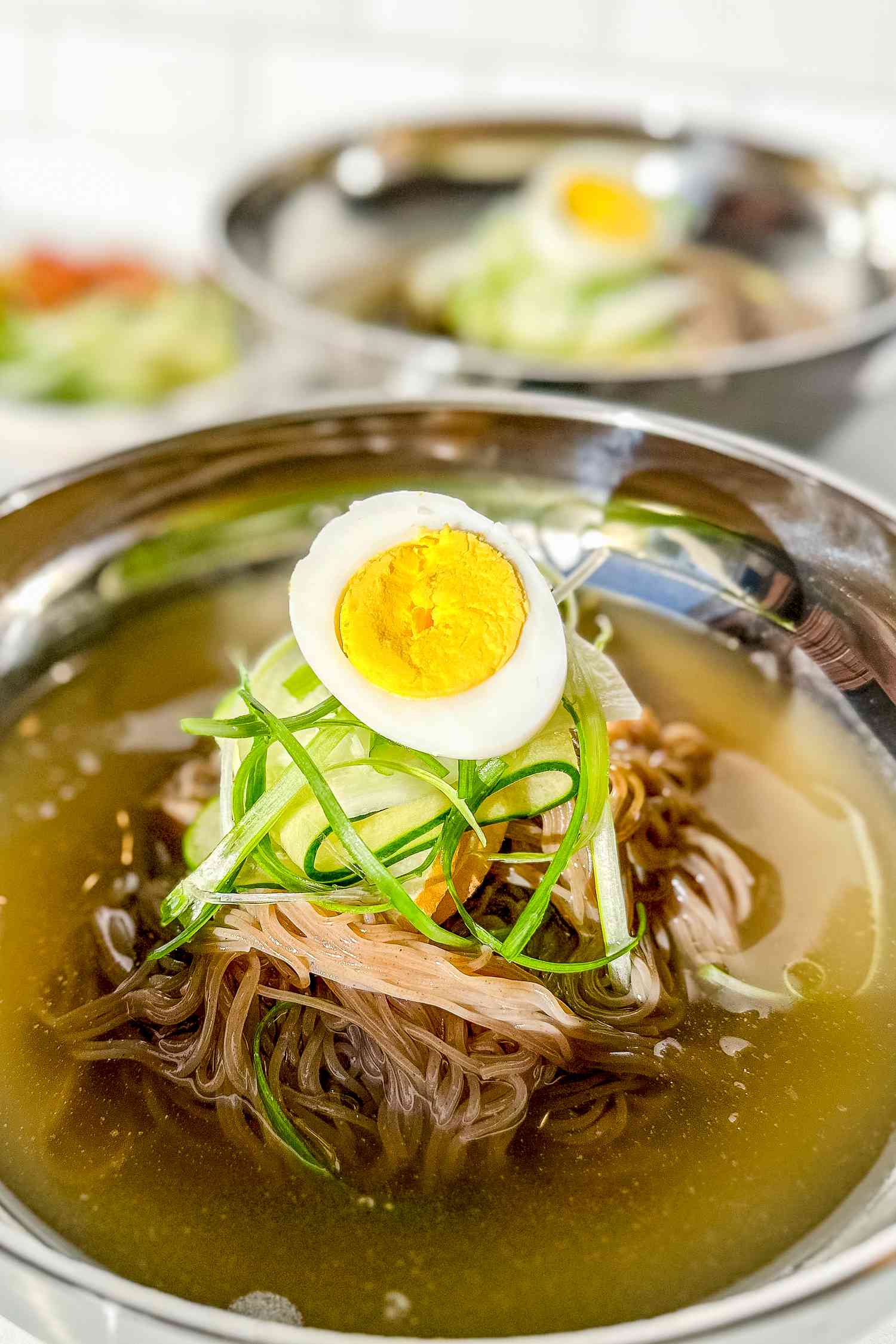 Bowl of Naengmyeon Topped With a Halved Hard Boiled Egg, Cucumber Ribbons, and Julienned Green Onions, and in the Background, Another Serving of Naengmyeon in a Bowl and Bowls Toppings