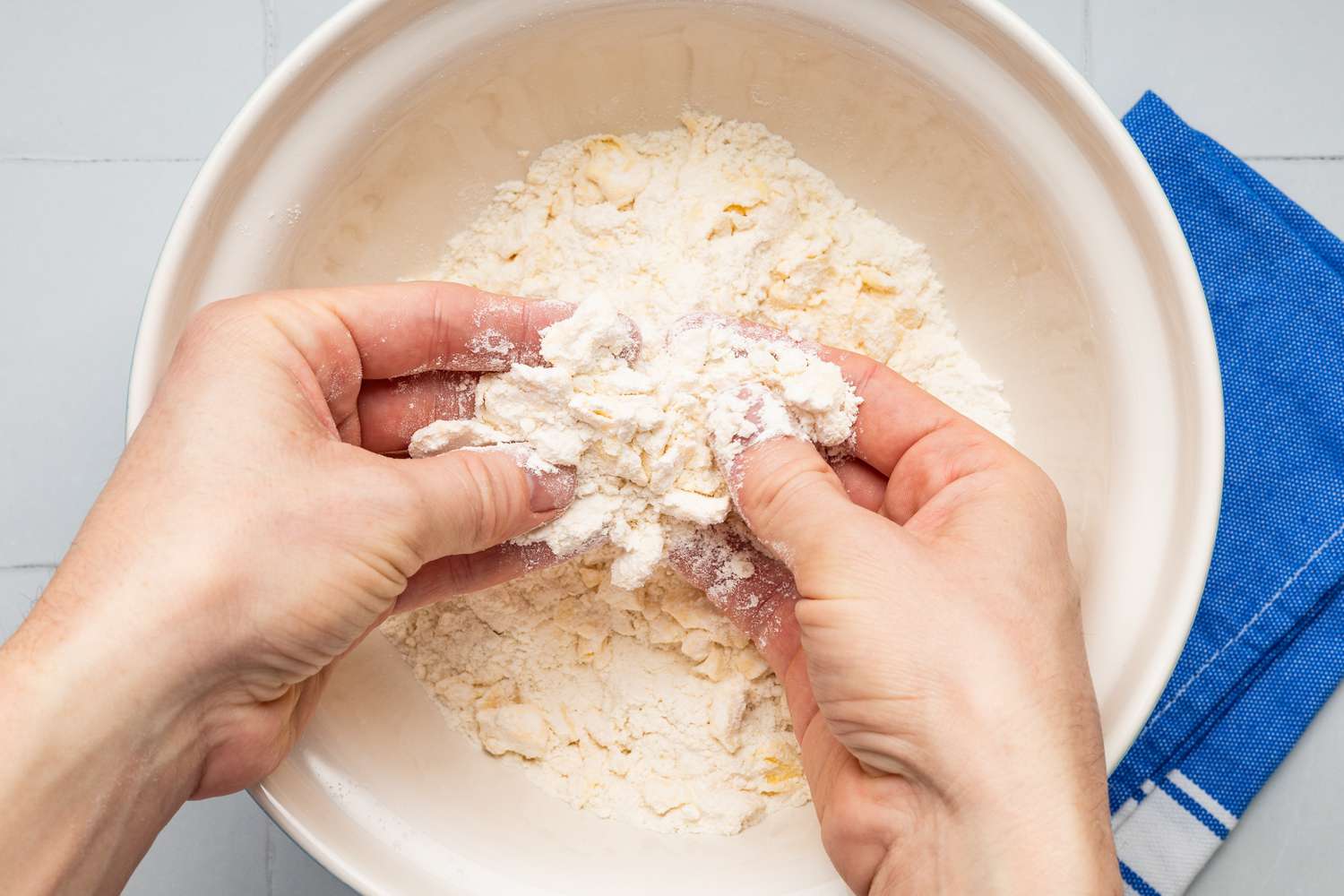 Hands breaking butter into smaller pieces while coating them in flour for drop biscuit recipe