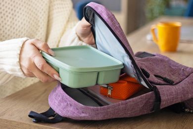 Woman wearing a white sweater putting a bento-style lunch box into a small purple backpack on a wooden tabletop
