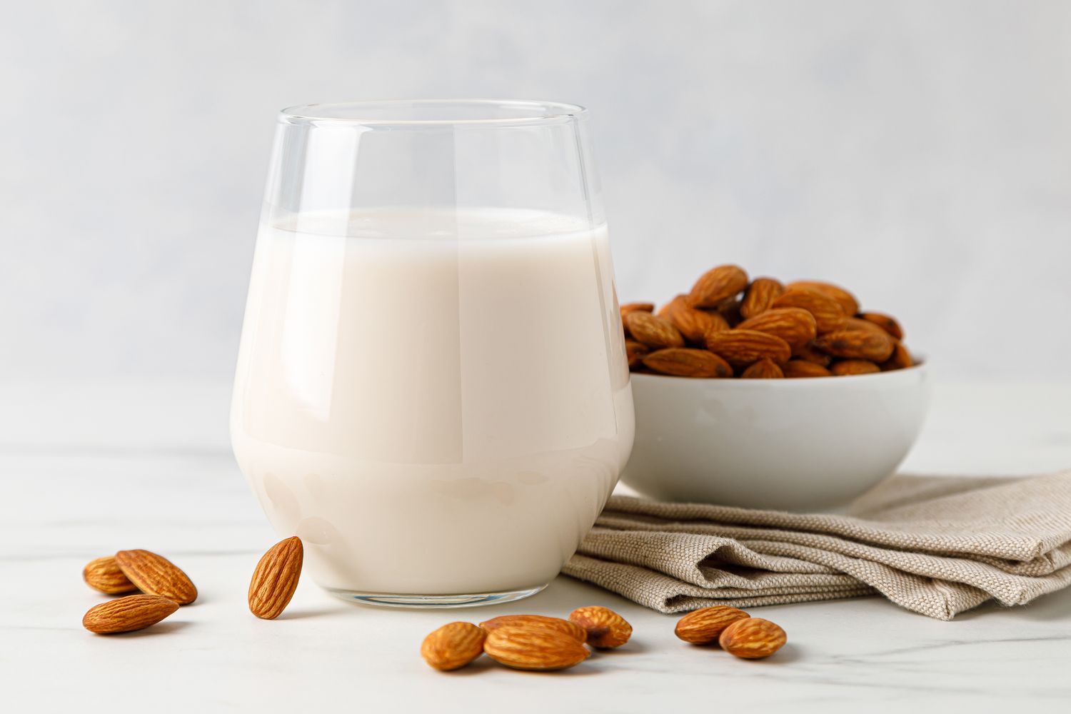 Side view of a glass of almond milk on a tabletop with a few almonds scattered around it next to a table napkin and a small bowl piled high with almonds