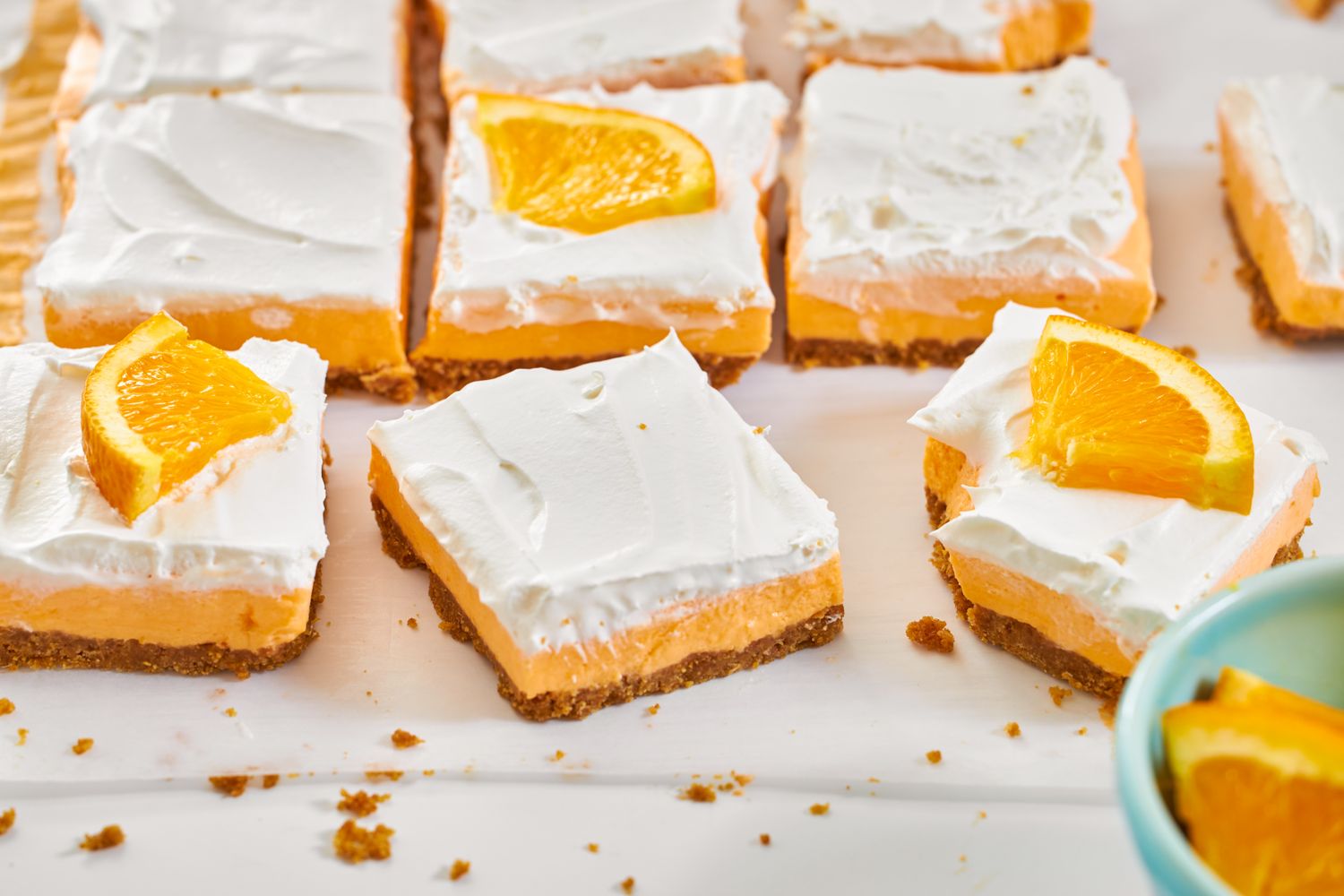 Creamsicle dessert bars with orange wedge topping and a graham cracker crust arranged on a white surface
