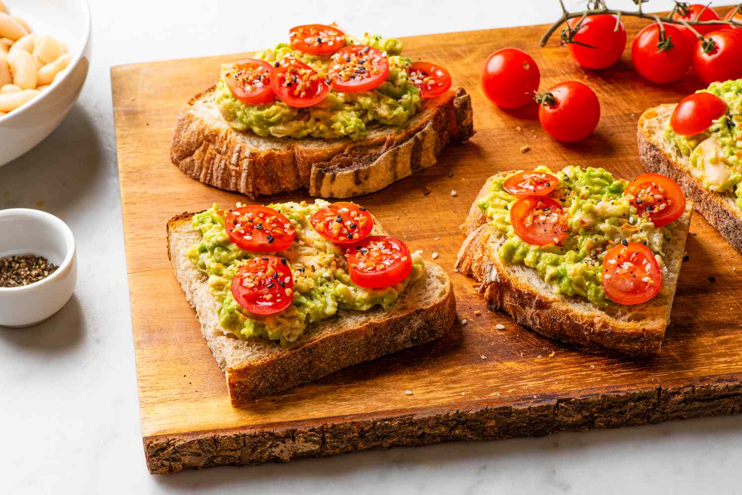 Avocado toast topped with tomato slices and seeds on a wooden board next to tomatoes and seasoning bowls