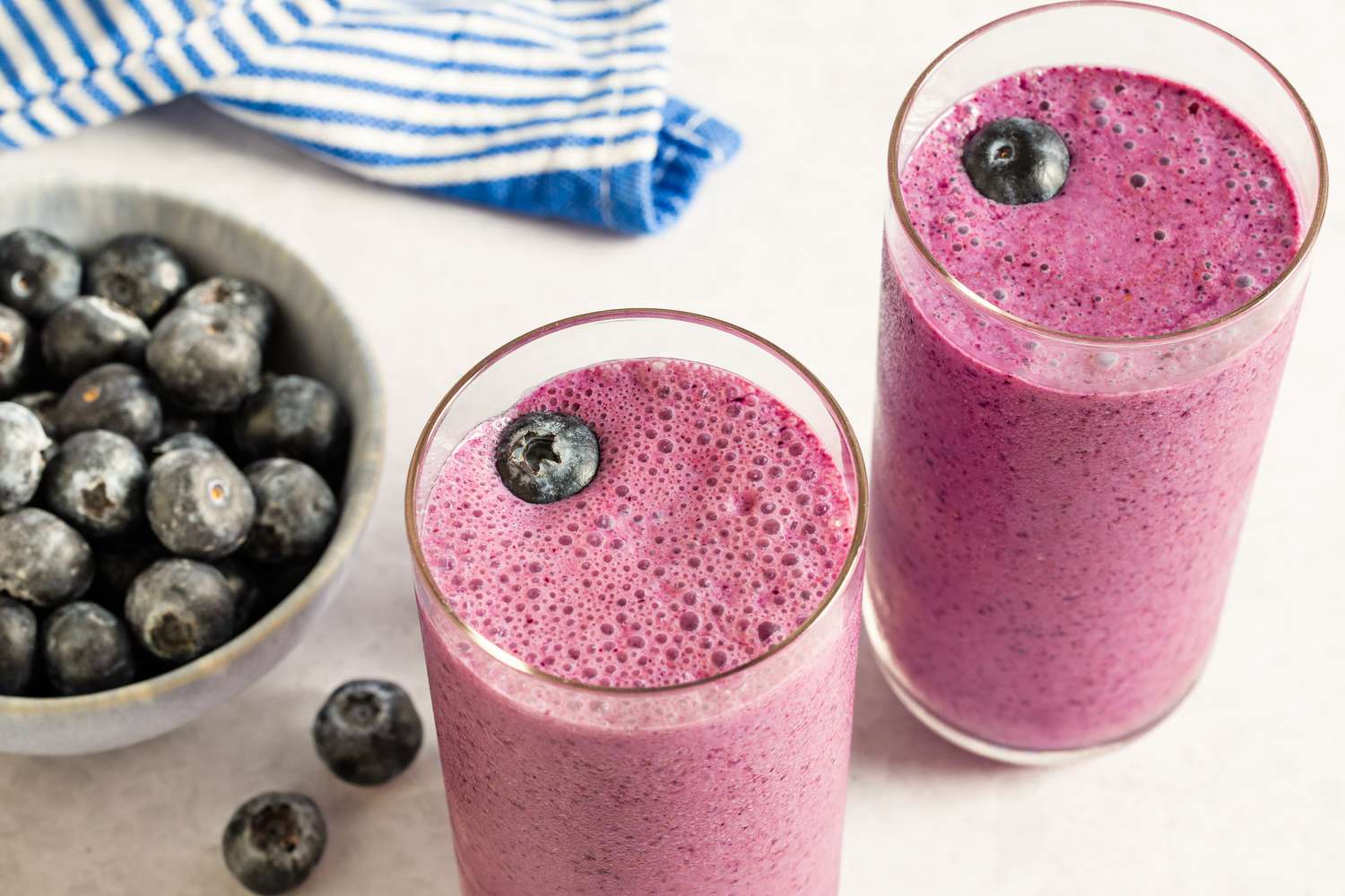 Two glasses of blueberry smoothie next to a bowl of fresh blueberries and a striped cloth