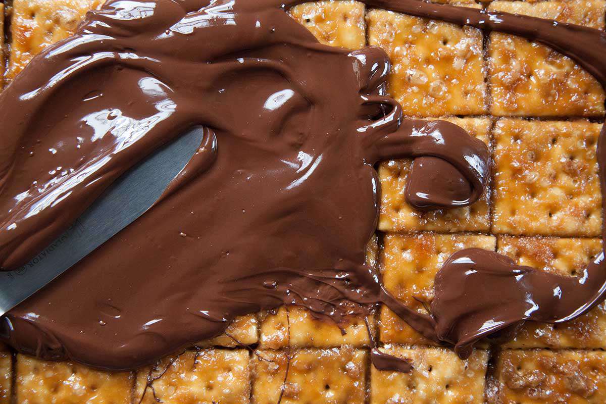Melted chocolate being poured and spread over the baked saltine crackers with a butter knife