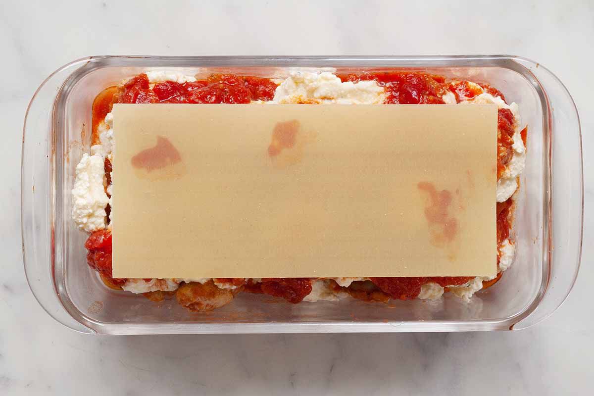 A glass loaf pan with uncooked noodle resting on top the layers of noodles, cheese and tomato sauce.