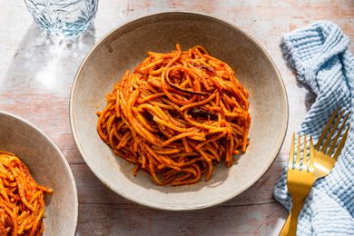 Bowlful of assassin's spaghetti at a table setting with more in a bowl, a glass of water, utensils, and light blue kitchen towel